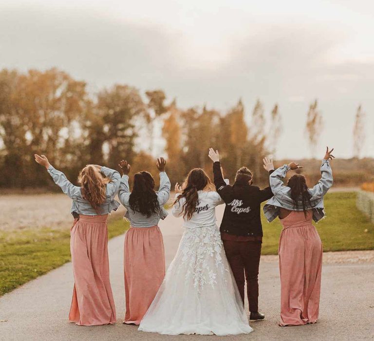 Bridal party wearing personalised denim jackets on top of dusky pink one shoulder bridesmaid dresses and illusion sleeve wedding dress and maroon bridal suit 