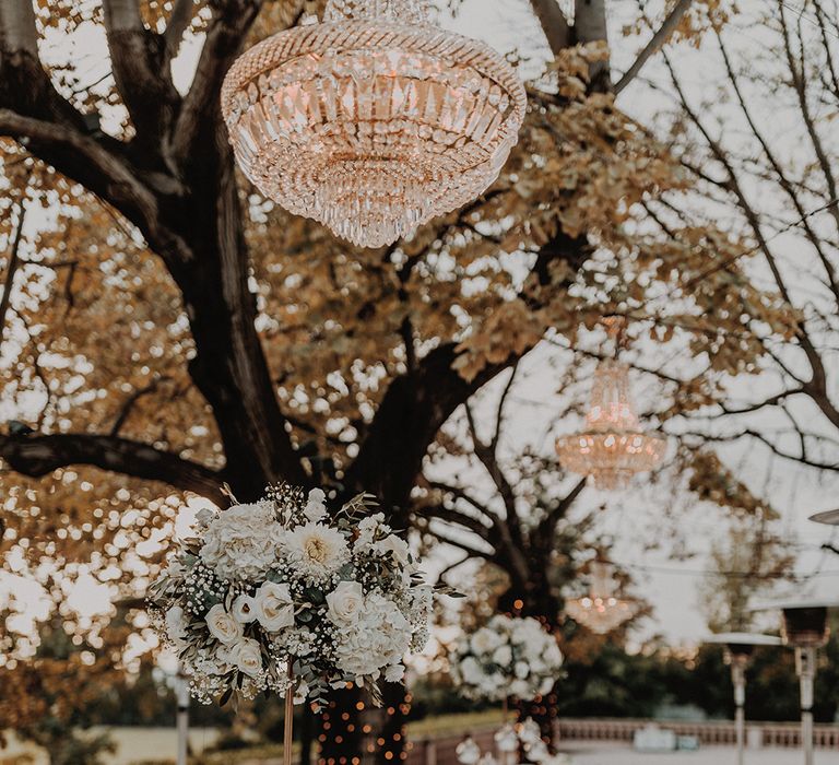 Outdoor wedding classic tablescape inspiration - white tablecloth, bejewelled outdoor chandeliers, fairy lights woven around the trees, peony and foliage table runners, and gold cutlery and crockery 