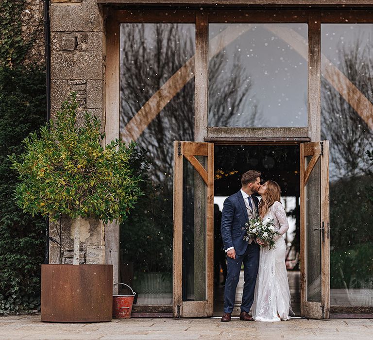 The bride and groom kiss at the entrance to Stone Barn as snow softly starts to fall 