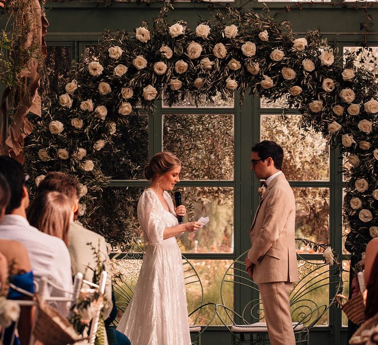 Bride in Grace Loves Lace bridal separates reading out her wedding vows to the groom standing in front of a white rose floral arch 