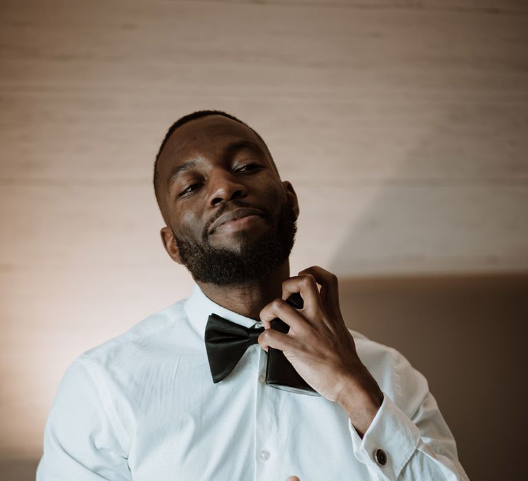 Black groom in a long sleeve white shirt putting on his black bow tie for the wedding 