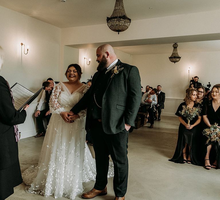 Groom standing at the altar in a green tweed wedding suit with the bride in a long sleeve lace wedding dress 