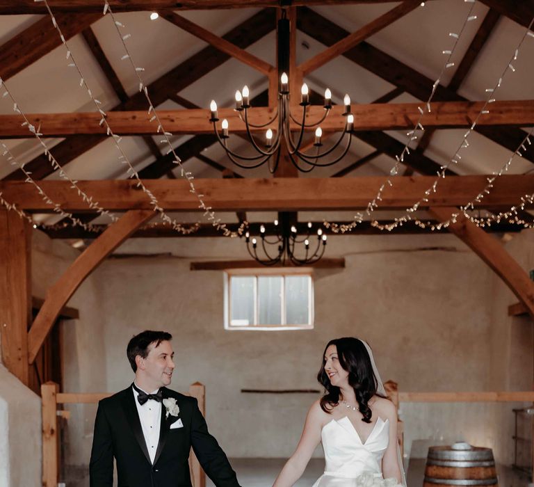 Bride in white silk wedding dress with a white rose bridal bouquet hols hand with groom in black tuxedo at Upton Barn wedding venue