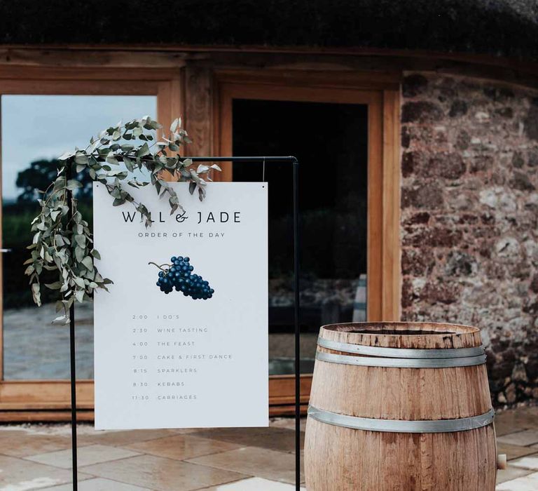 Minimalistic white wedding welcome sign with grape illustrations on black copper frame with eucalyptus decorations at Brickhouse Vineyard Devon wedding