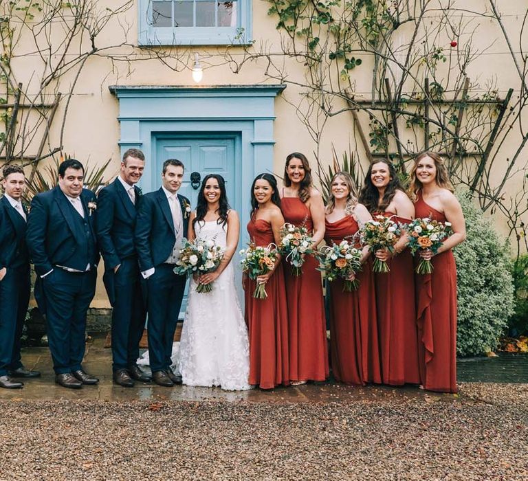 Groom in navy grooms morning suit with grey waistcoat, off-white tie and mixed wildflower boutonniere standing with groomsmen in matching groomsmen morning suits and bride in a-line tulle wedding dress with beaded lace applique with petals and a square neckline with side tulle cutouts standing with bridesmaids in cinnamon mismatched bridesmaid dresses 