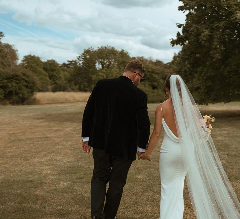 Bride in low back satin wedding dress with button back detail with pearl wedding veil walking along with groom in black tie 