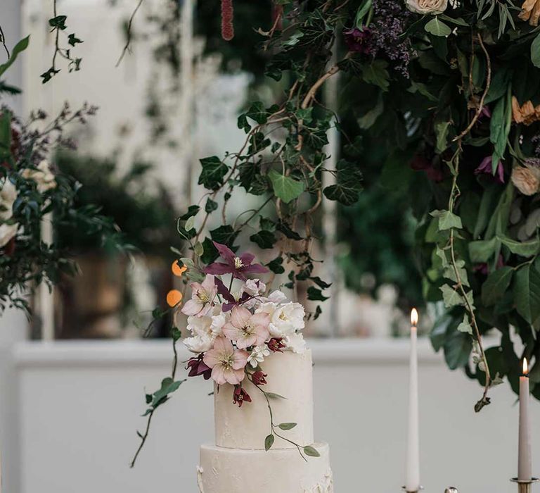 Four tier white wedding cake with intricate icing details and dried flower wedding cake topper on wooden dessert table with wedding cupcakes and wedding biscuits with twisted tapered candles in neutral shades in gold candlesticks surrounded by wildflower and foliage wedding flower arrangements