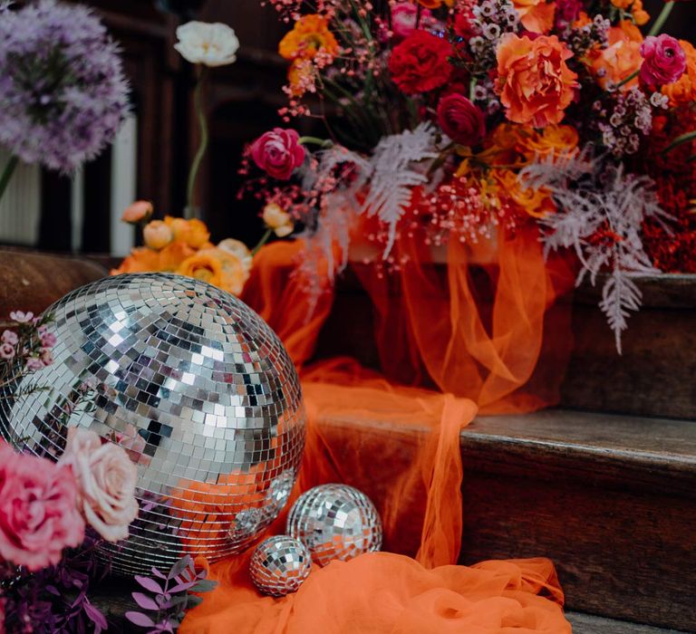 Large disco ball wedding decor on orange tulle fabric and colourful flower arrangements with pink garden roses, purple giant alliums, dark red poppies and foliage 