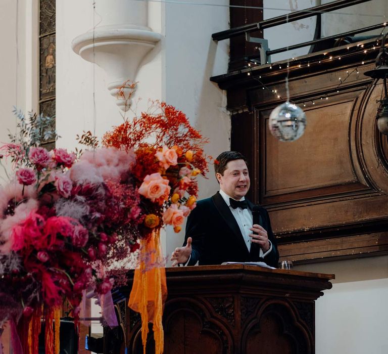 Groom in velvet tuxedo and black bowtie standing at the podium of Fabrica Brighton wedding venue doing grooms speech surrounded by suspended colourful flower arrangements and suspended disco ball wedding decor