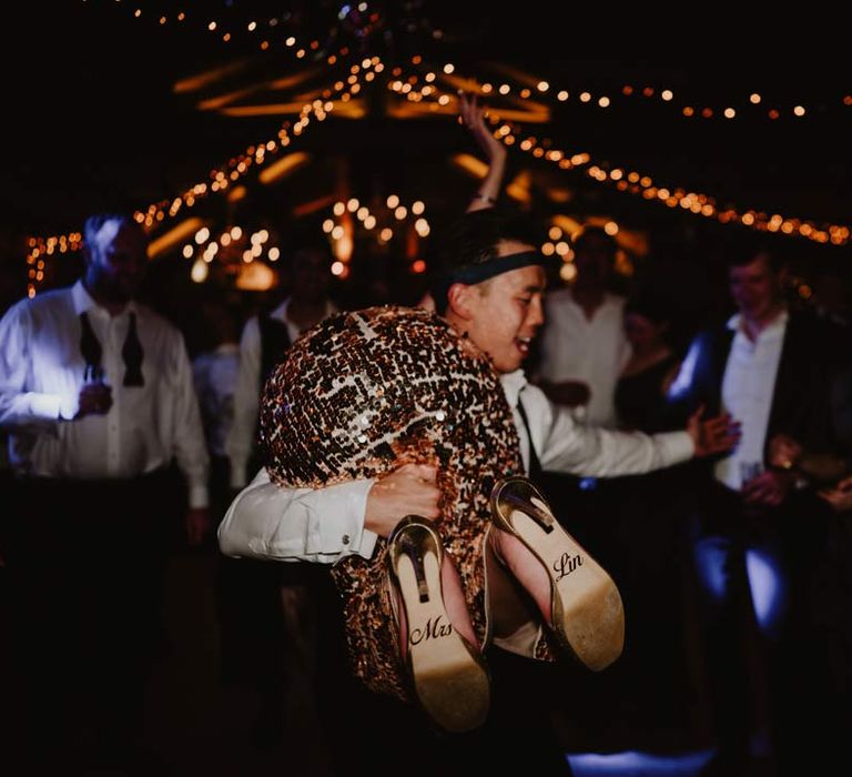 Groom in white shirt with black tie around his head holding up bride in gold sequin sparkly reception dress over his shoulder