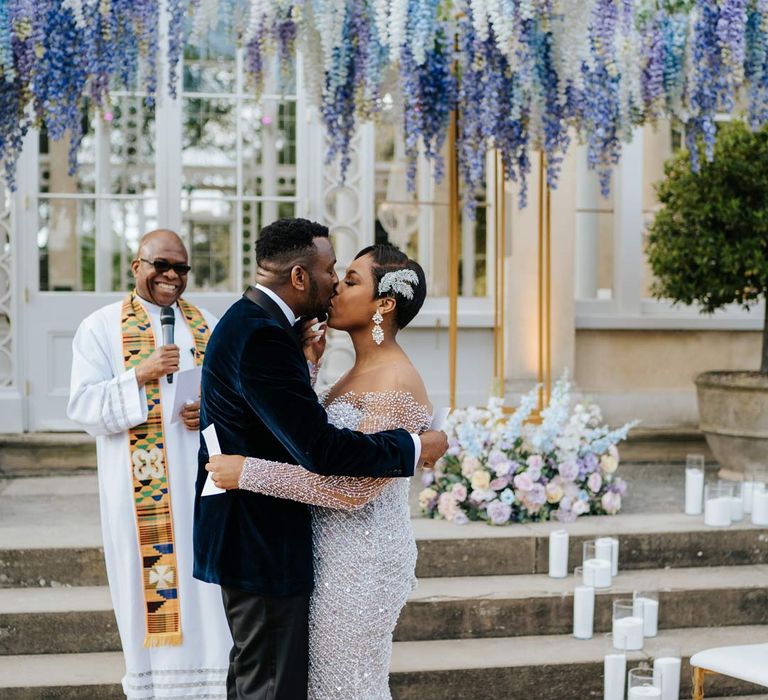 Groom in velvet midnight blue double breasted groom’s blazer, velvet bowtie and peach garden rose boutonniere kissing bride in illusion v-neck sheer off shoulder long sleeve pearl embellished wedding gown standing underneath suspended pastel flower arrangements at Syon Park vow renewal 