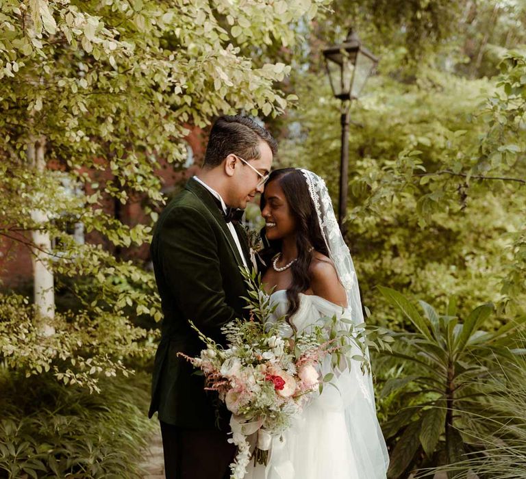 Bride in off the shoulder layered tulle wedding dress and floral embroidered cathedral-length veil embracing groom in bottle green velvet grooms suit, black bowtie and chic boutonniere on the grounds of Inner Temple Hall