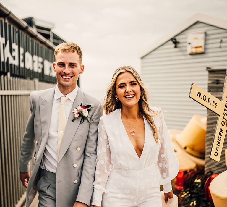 Groom in a light grey wedding suit holding hands with his bride in a long sleeve wedding jumpsuit at East Quay coastal wedding 