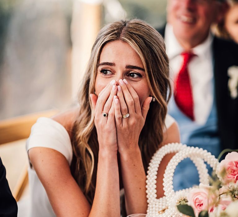 bride with her hands over her mouth in shock and a pearl handbag in front of her during the marquee wedding reception speeches 