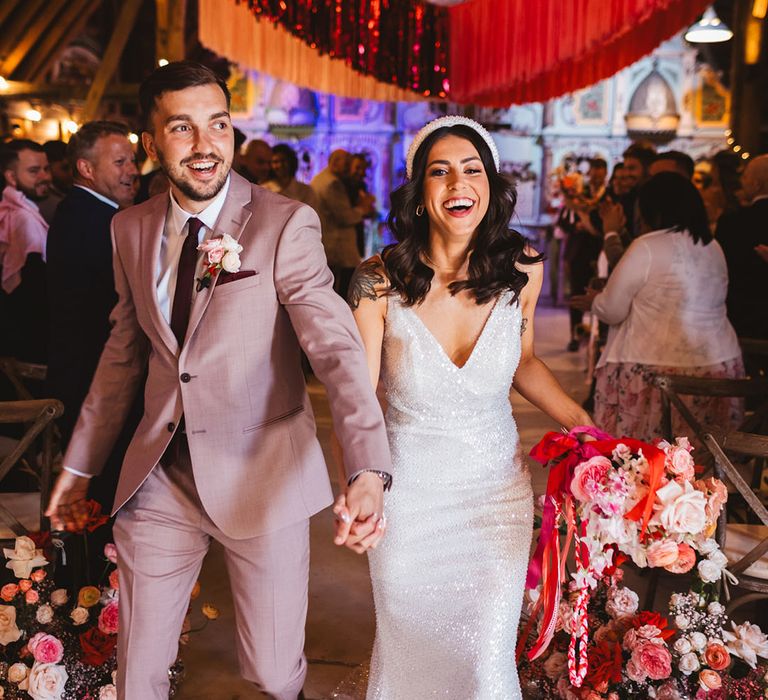 Bride wearing sparkly Made With Love wedding dress with headband walking with groom in taupe suit under colourful streamer decorations