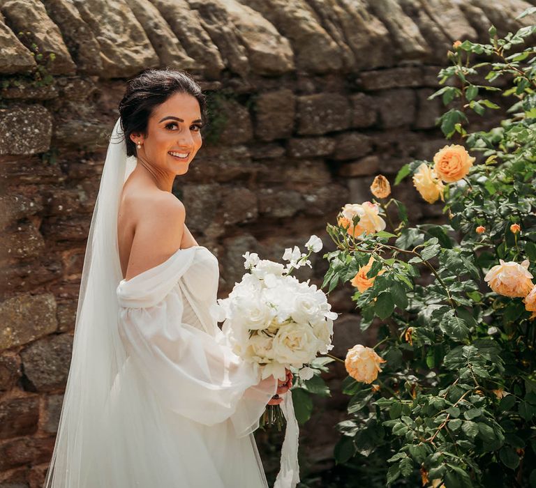 Bride in off-the-shoulder white tulle wedding dress hold white rose wedding bouquet with hair in a bridal updo and veil