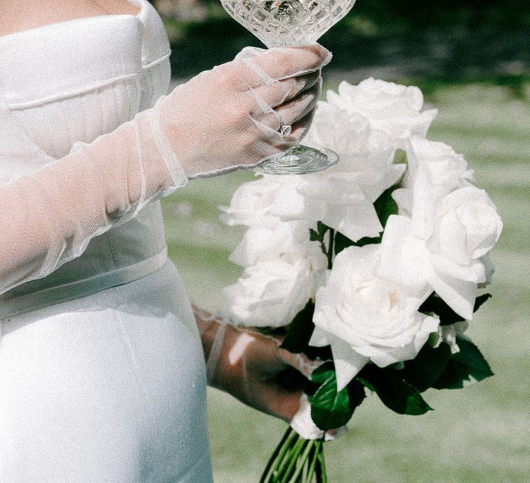 Bride with tulle sheer bridal gloves holding a champagne coupe and white rose long stem wedding bouquet 