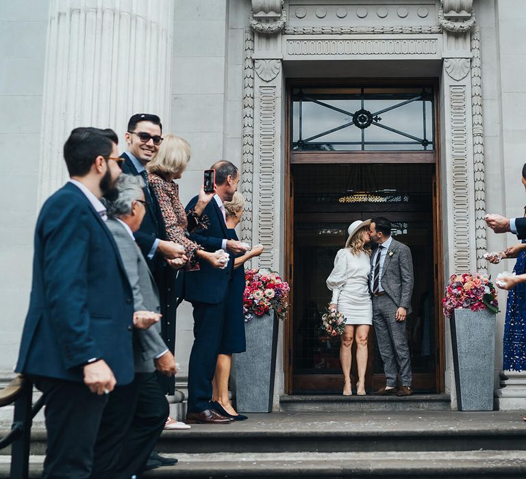 Bride and groom kissing outside wedding venue with guests waiting for confetti moment at intimate London wedding