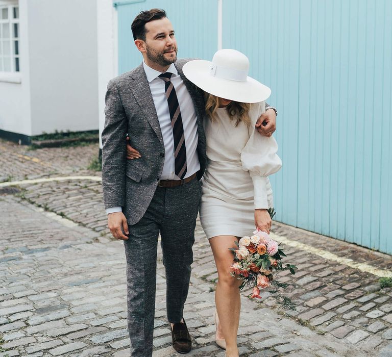 groom holding his new bride wearing grey suit and brown and black stripe tie bride wears short Net A Porter wedding dress with white hat