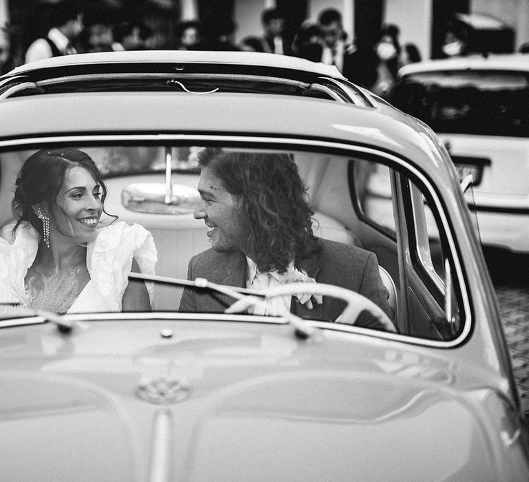 Bride with chandelier earrings and ruffle sleeves and groom with curly hair sitting in their wedding car 