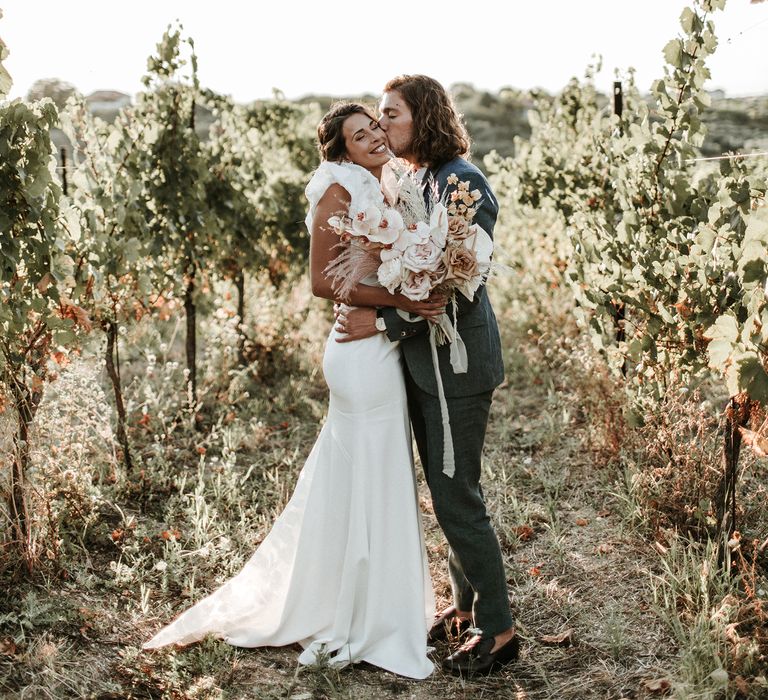 Bride and groom kissing in a vineyard with the bride holding an orchid wedding bouquet 