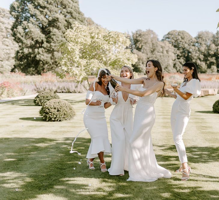 Bride and bridesmaids in all white dresses popping champagne on the lawn at Middleton Lodge gardens 