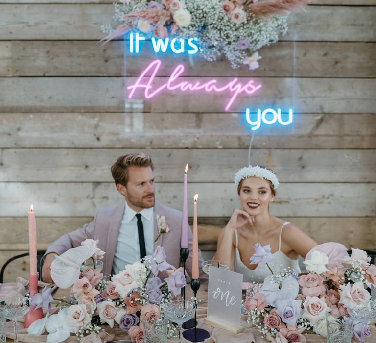 Bride and groom sitting at an intimate table with a 'It was always you' pink and blue neon sign with flower cloud backdrop