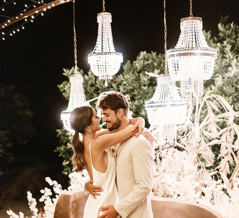 Bride in a satin slip dress kissing her groom in a beige suit under a canopy of chandelier lights 