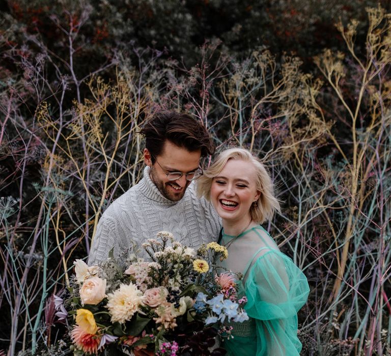 Bride and groom laughing at their beach elopement standing infant of a sand dune with colourful flower stems 