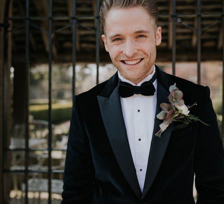 Smiling groom in black velvet Hugo Boss suit and black bow tie with white rose buttonhole