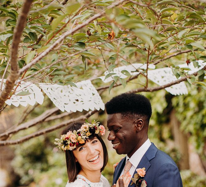 Groom in a navy blue suit embracing his bride in a Broderie Anglaise wedding dress under a canopy of laser cut bunting