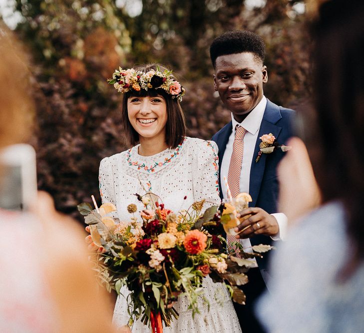 Bride and groom smiling during the drinks reception with the bride wearing a colourful flower crown and holding a wildflower bouquet 