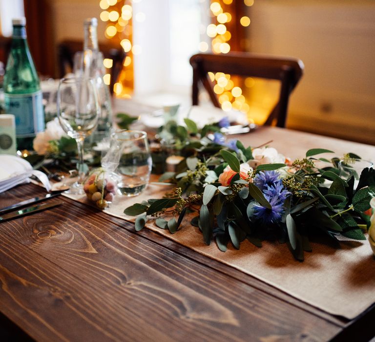 Florals run through the middle of wooden table with fairy lights in the background