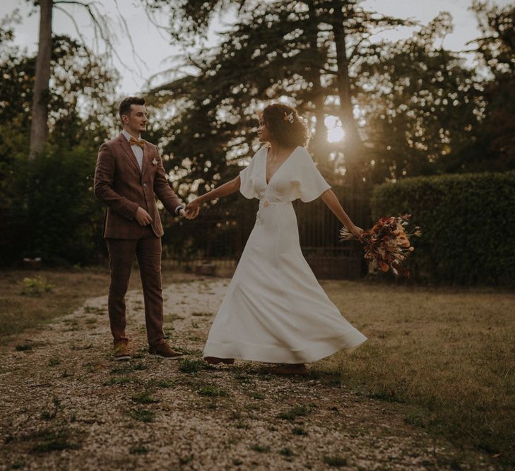 Groom in a brown wedding suit twirling his bride during golden hour in a minimalist wedding dress 