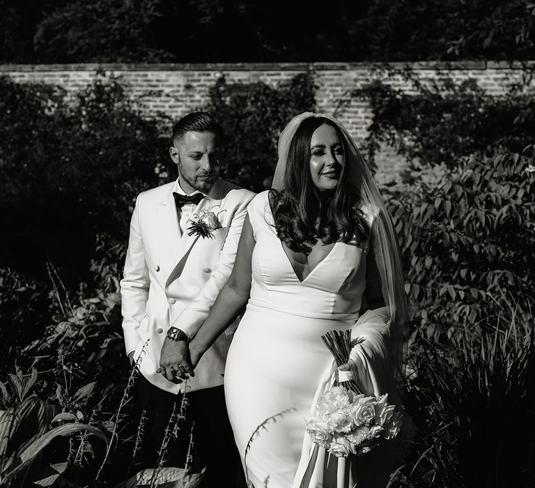 Bride in V neck Made With Love wedding dress and veil holding white rose bouquet walks outside with groom in white double breasted tuxedo jacket