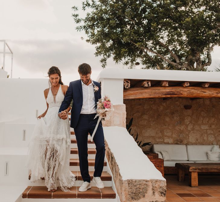 Groom in a navy suit and white trainers holding his bride's hand in a bohemian wedding dress with tassels 