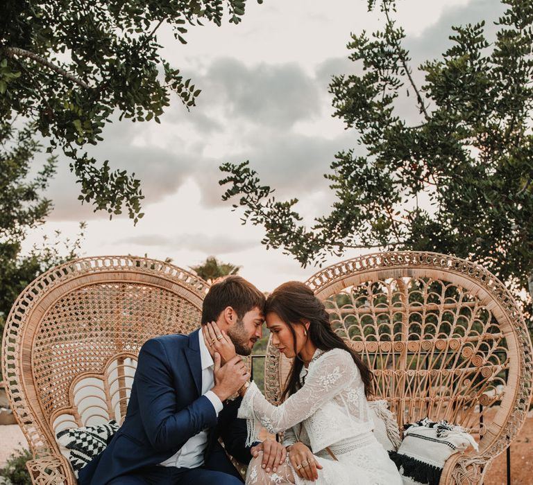 Bride in a Marylise Bridal gown with lace detail sitting on a peacock chair with her groom in a navy suit 