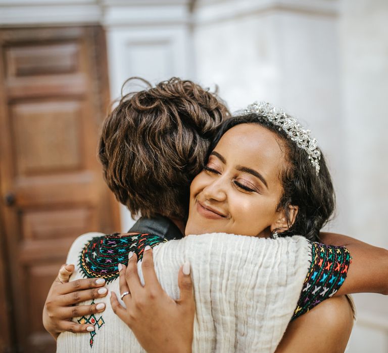 Bride with shimmering eye shadow, long eyelashes and beige nail polish hugging a wedding guest 