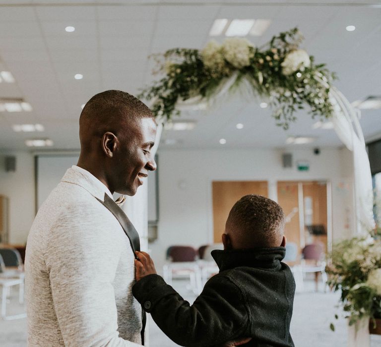 Groom in grey suit jacket with black lapels smiles as young wedding guest in black jacket holds on to his front at Bridge Community Church wedding