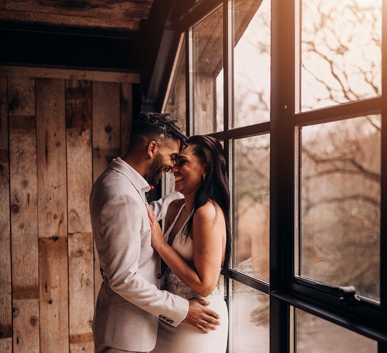 Bride & groom stand beside window as they look lovingly at one another 