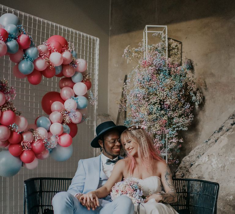 Bride and groom sitting on a black wire love seat surrounded by pastel gypsophila flower clouds and balloon installations