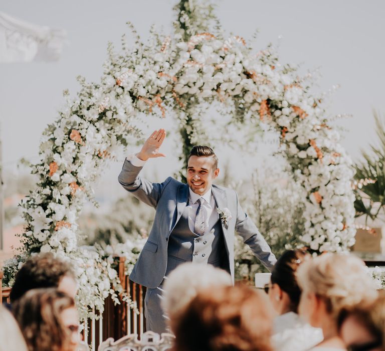 The groom waving to the guests as he waits at the altar under a white flower arch