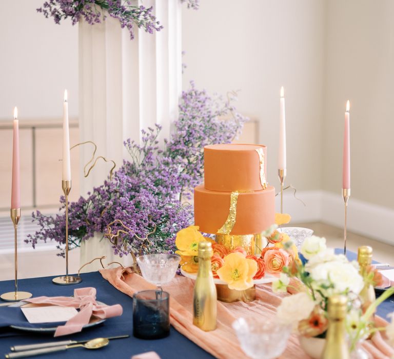 Orange frosted wedding cake complete with gold leaf and floral decor next to lavender display winding up pillar