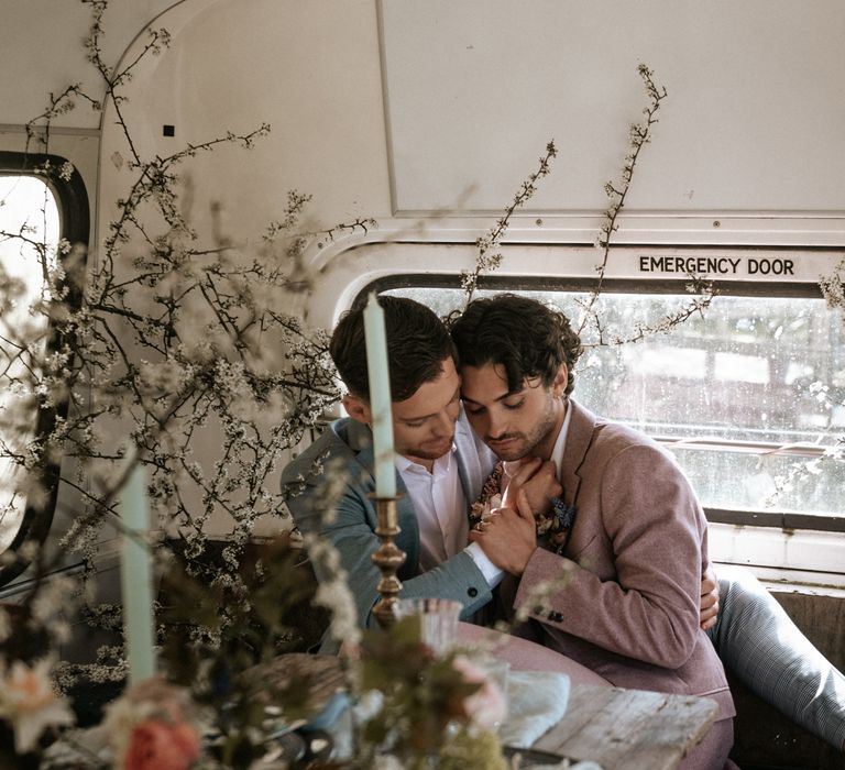 Grooms sit closely with one another on their wedding day whilst wearing pastel suits