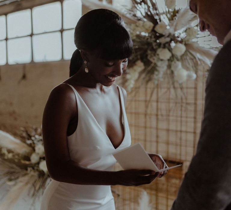 A bride and groom stand in front of a metal frame structure and she reads her vows from a book.