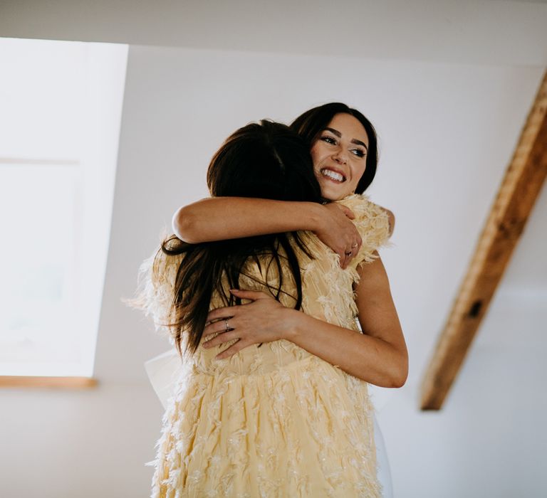 Bride in white Rebecca Vallance Dress hugs guest in yellow short sleeved dress with feather detailing before Harrogate wedding