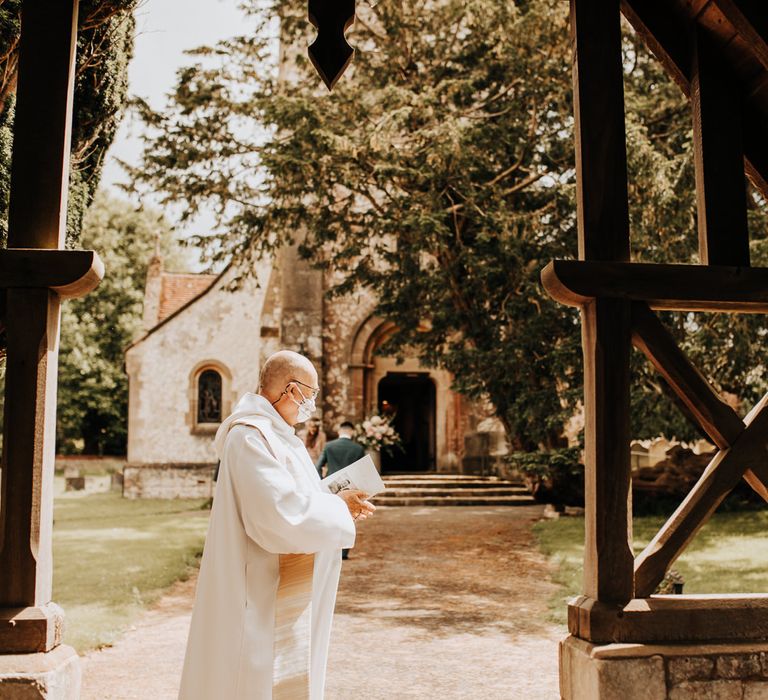 Vicar in white robes and mask stands in sunlit churchyard before wedding