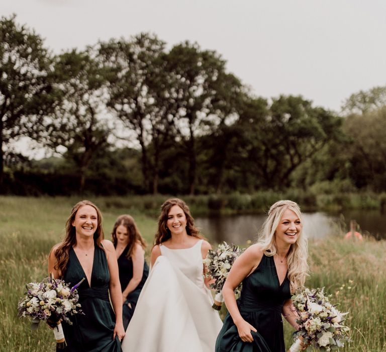 Bride in white Elbeth Gillis wedding dress and white wellies walks with three bridesmaids in first green v neck bridesmaids dresses and black wellies by pond at home farm wedding