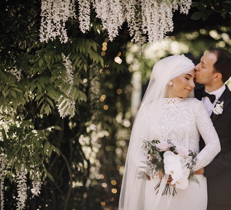 Groom kisses bride's forehead under hanging wisteria with bridal hijab and veil