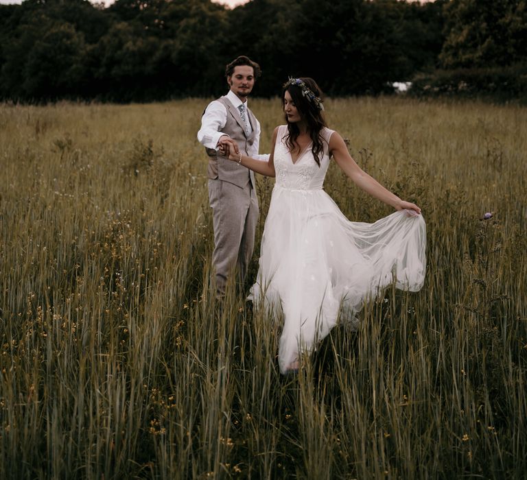 Bride & groom walk through green fields with one another as groom holds her hand